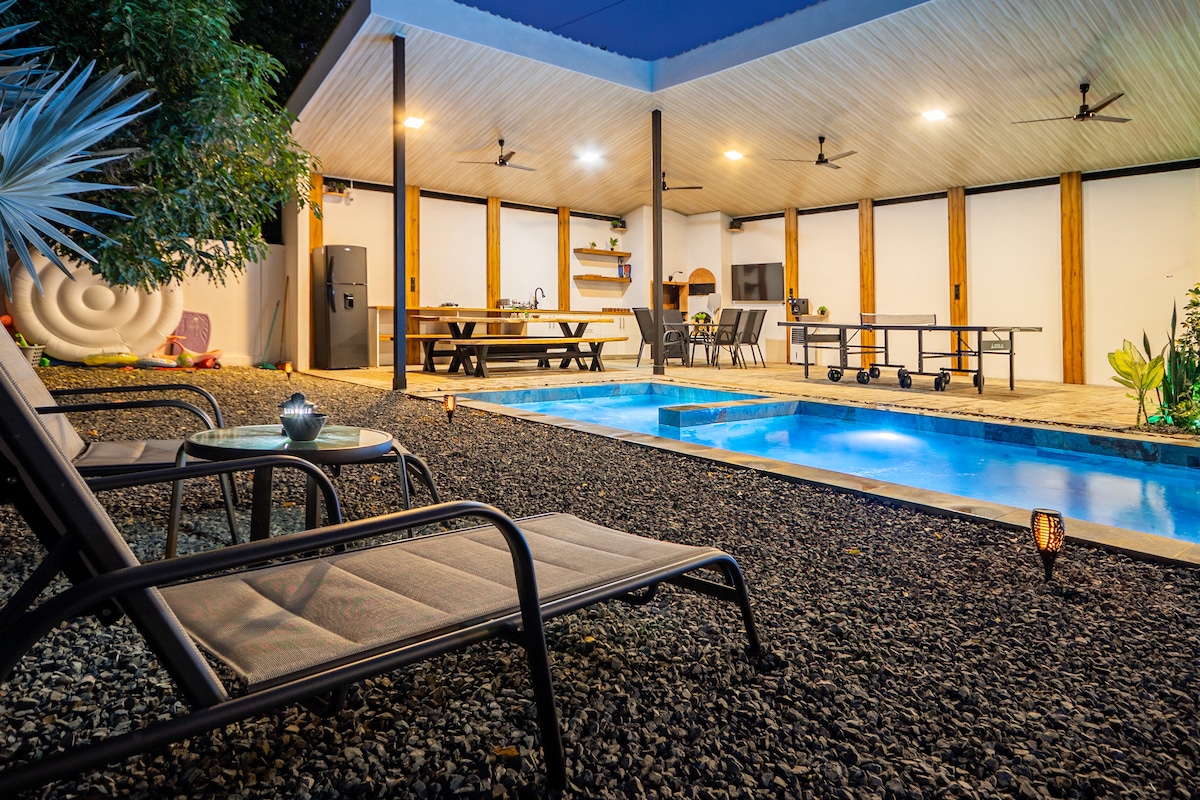 A spacious outdoor area features a large saltwater pool surrounded by lounge chairs. An outdoor dining space and game table are visible under a covered patio, marked by ceiling fans and natural wood accents. Lush greenery enhances the tranquil setting.