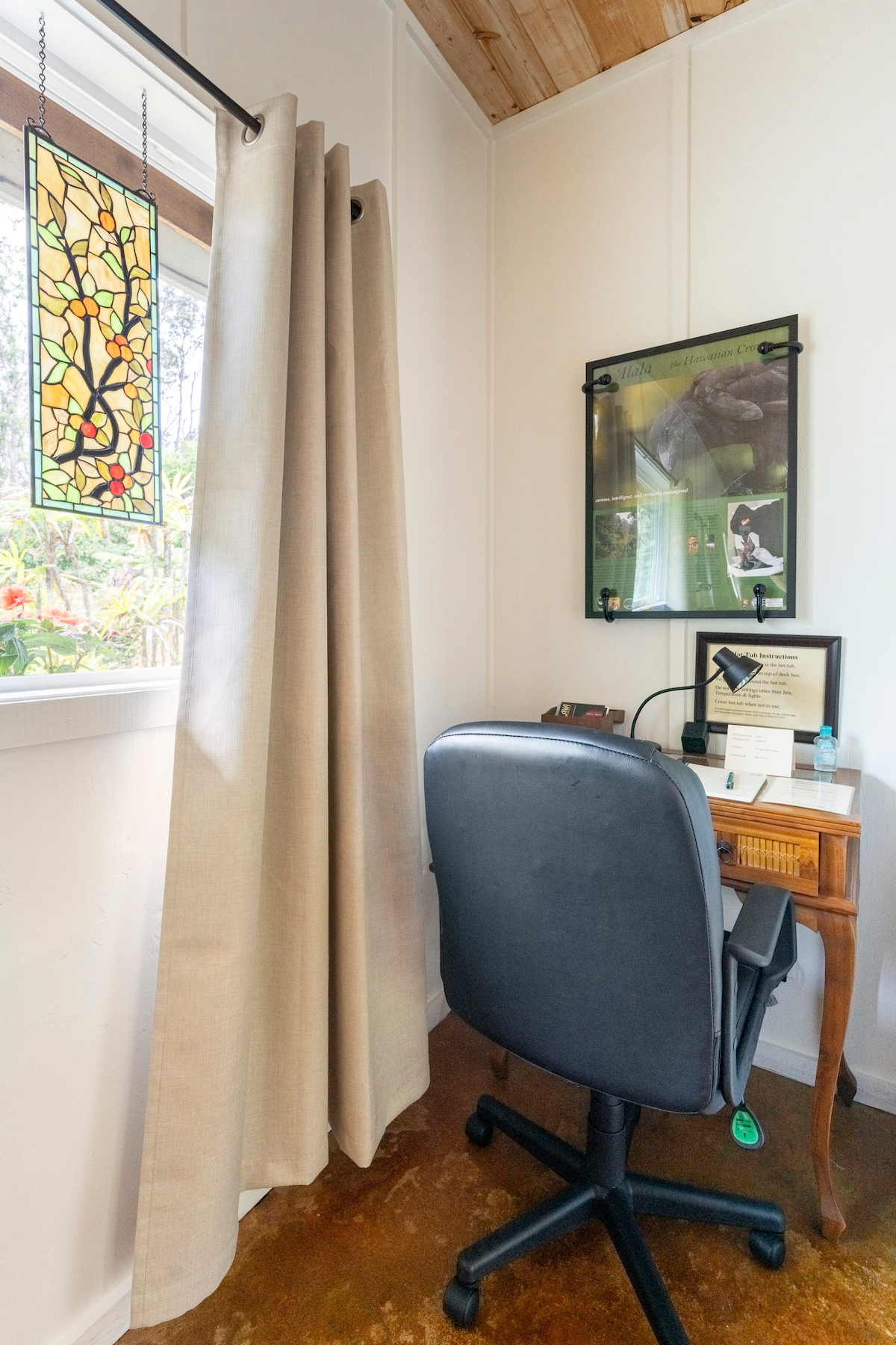 A small workspace is situated near a window, featuring a wooden desk and a comfortable office chair. A stained glass panel adds color to the scene, while a framed piece of art is visible above the desk. Natural light illuminates the area.