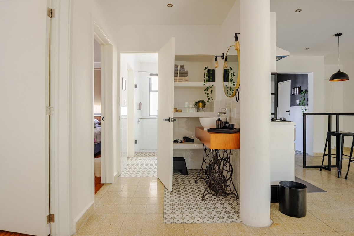 An open kitchen space showcases a modern design with a wooden countertop featuring a stylish sink, complemented by greenery. A high table with two stools can be seen in the foreground. Tiled flooring adds texture, while doorways lead to nearby rooms.