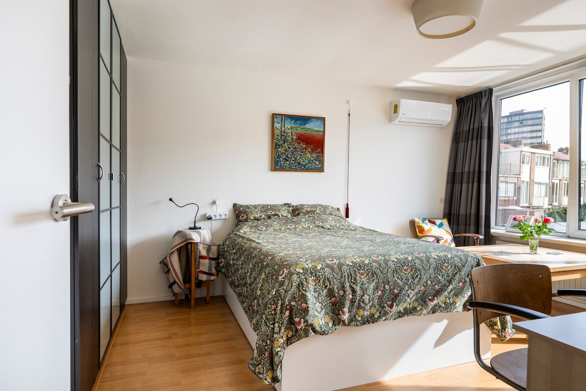 A cozy sleeping area features a spacious bed adorned with a floral-patterned cover. A small desk and chair are positioned near the window, which allows natural light to brighten the room. A piece of artwork hangs on the wall, and a plant adds a touch of freshness.
