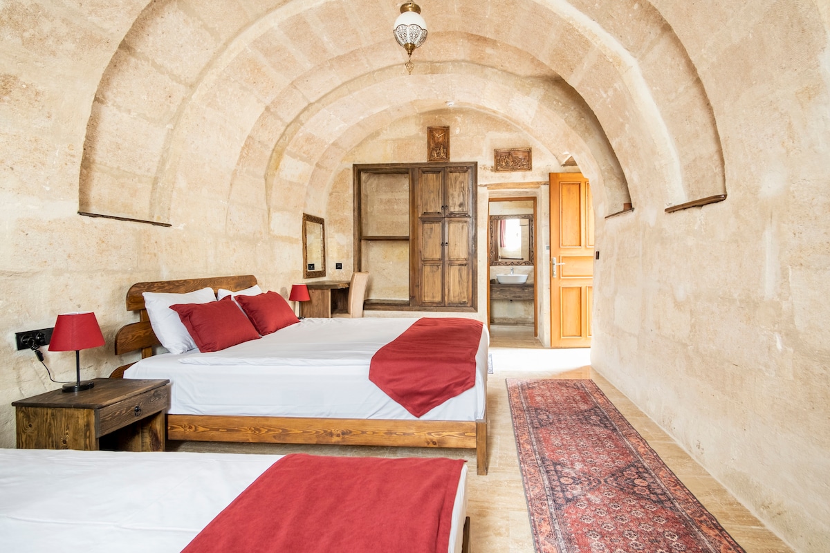 A room with curved stone ceilings contains two wooden beds dressed in white linens with red accents. A traditional area rug adds color to the wooden floor. Visible wooden doors lead to an adjoining bathroom, enhancing the room's spacious layout.