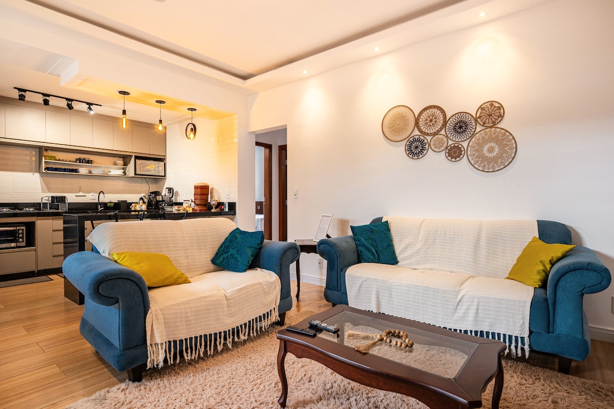 A comfortable living area features two blue sofas draped with white throws, complemented by decorative pillows. A wooden coffee table sits at the center on a soft area rug. The open-plan kitchen is visible in the background, showcasing modern appliances and a bright atmosphere.