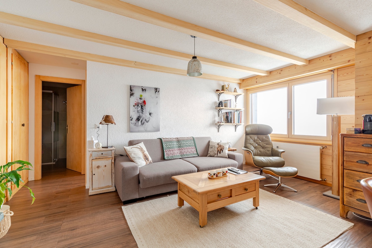 A cozy living space features a large gray sofa alongside a comfortable armchair. Light wood beams stretch across the ceiling. A wooden coffee table is placed on a neutral rug, and shelves display books. Windows provide natural light and views of the surroundings.