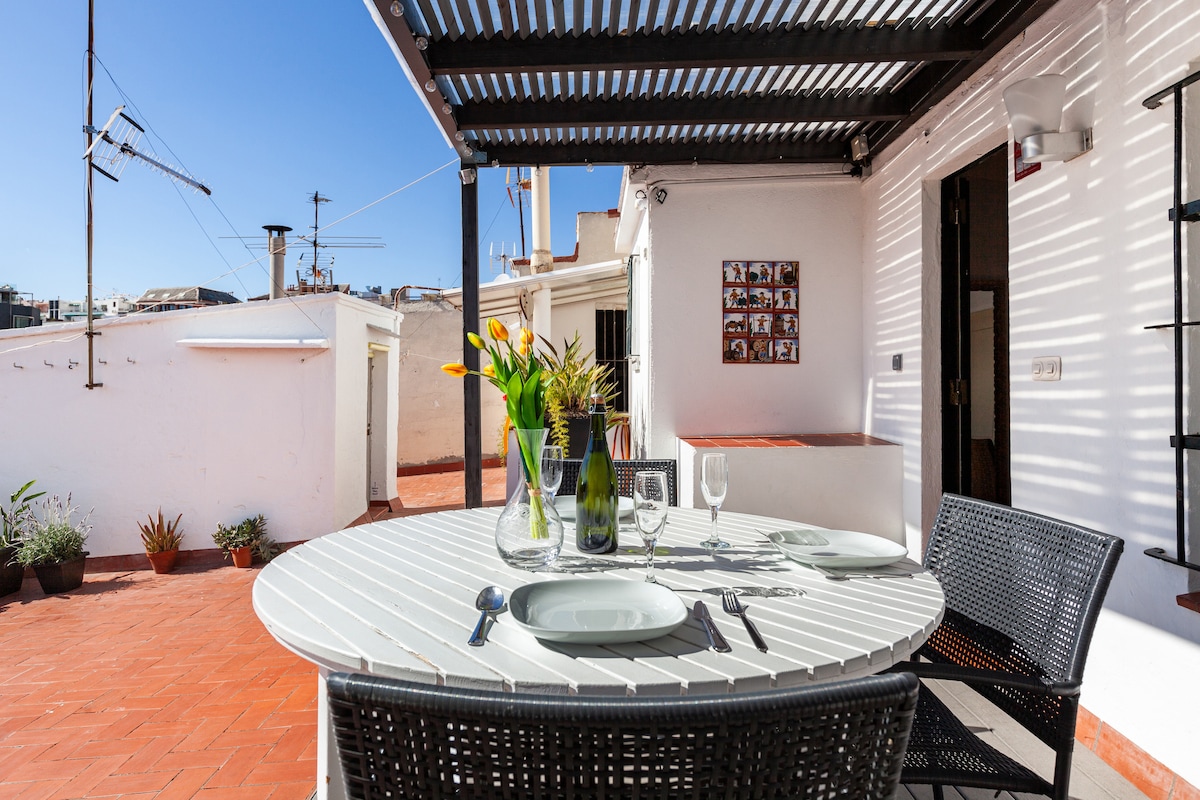 An outdoor dining area is presented under a slatted pergola, featuring a round table set for four. Vases containing colorful flowers are noticed, alongside potted plants around the terrace. Sunlight illuminates the space, highlighting views of the surrounding rooftops.