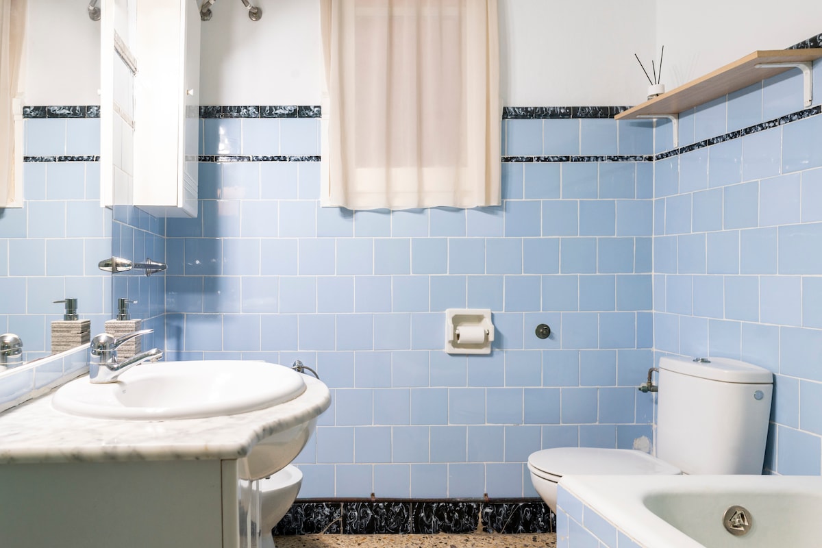A tiled bathroom features light blue walls and a marble countertop. A sink, toilet, and a small bathtub are present. Natural light filters through a window with sheer curtains, providing a bright and airy feel. Shelving above the sink offers space for toiletries.