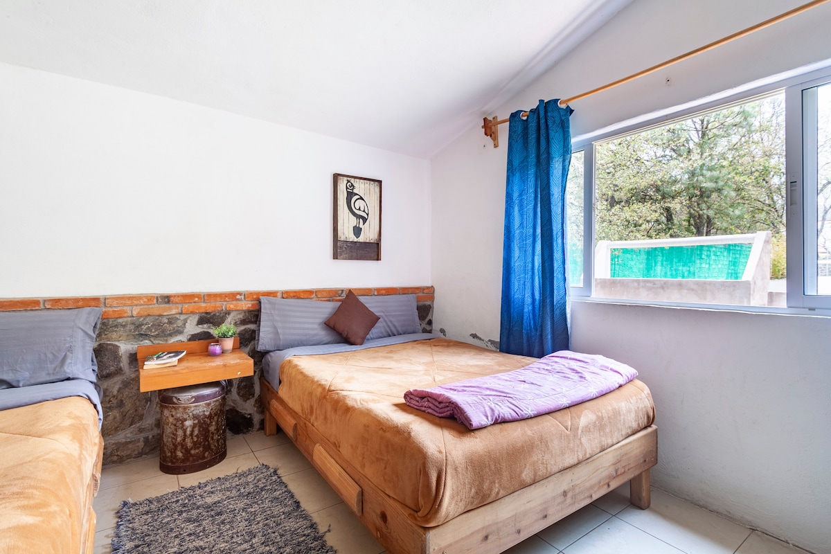 A serene sleeping area features two spacious beds dressed in warm, earthy tones. Natural light enters through a large window adorned with sheer blue curtains, illuminating the room. A small wooden table completes the space, providing an inviting atmosphere for relaxation.