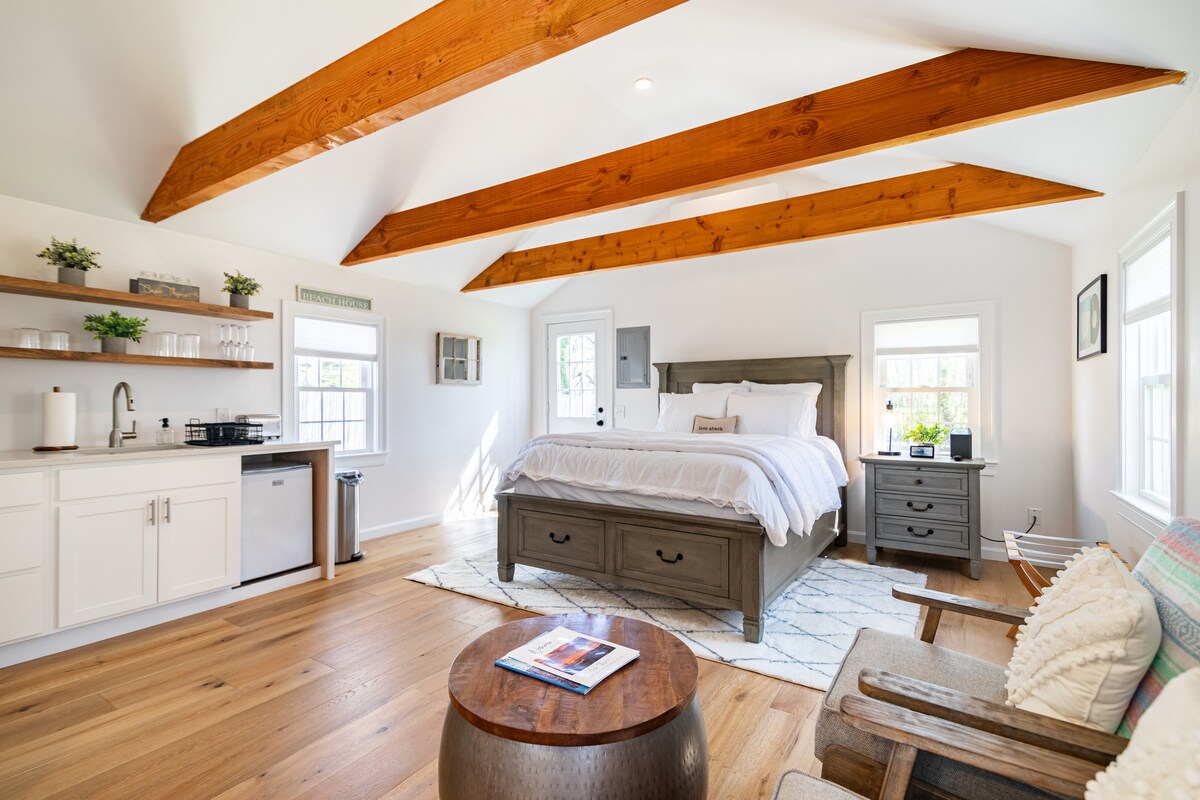 An airy studio space features high-beamed ceilings that enhance its open feel. A spacious bed is centered in the room, backed by a large window, while natural light illuminates the wood flooring. The kitchenette, along with seating areas, provides functionality and comfort.