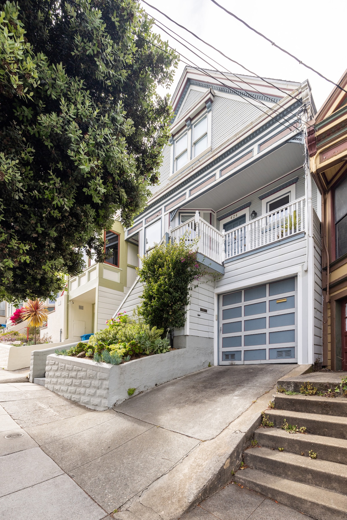 A charming cottage is viewed from the street, showcasing a two-story structure with a light-colored facade and balcony. Green foliage is present at the base, and the driveway features a garage door. The sloped pedestrian path leads to steps ascending the side.