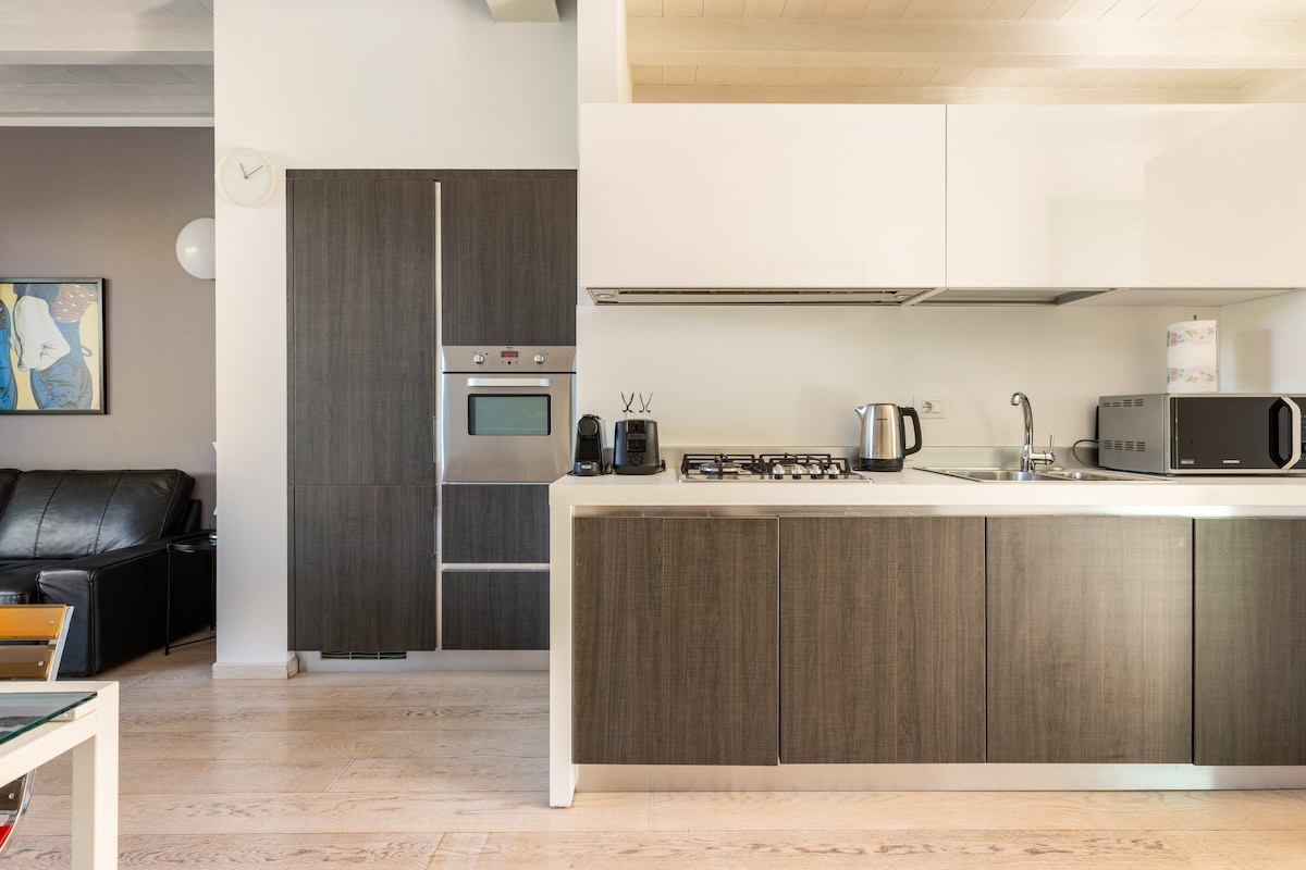 A modern kitchen is displayed, featuring sleek cabinetry and stainless steel appliances. A built-in oven and stovetop are visible along with essential kitchen items, including a kettle and microwave. Light wood flooring complements the contemporary design.