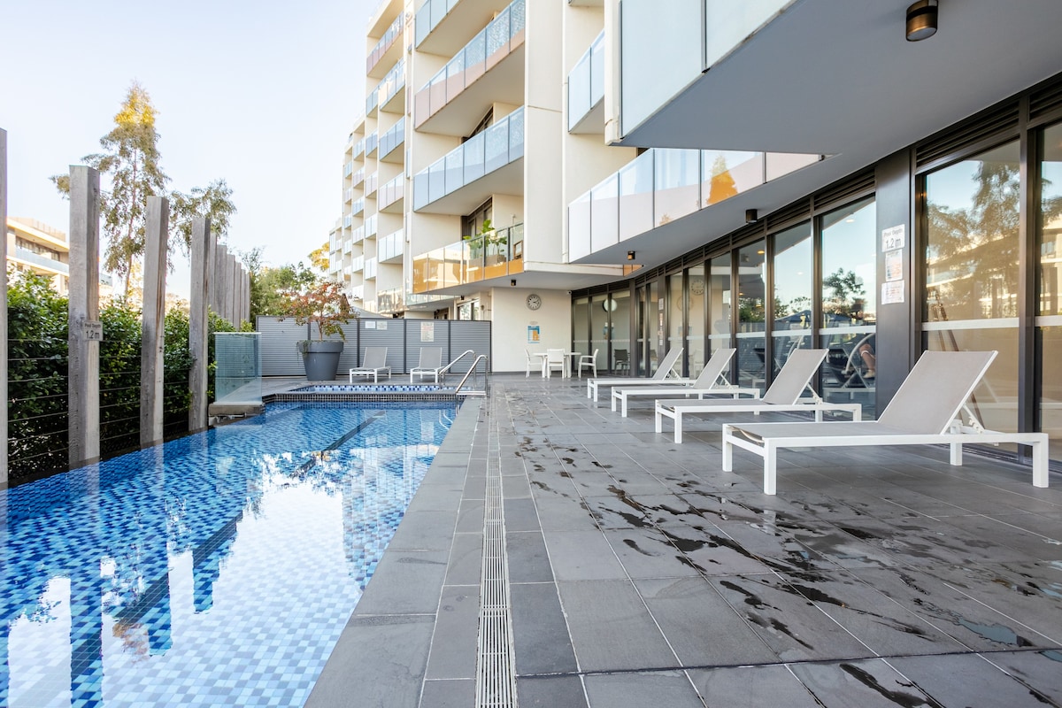 A modern pool area is presented, featuring a shimmering blue tiled pool surrounded by sleek grey stone flooring. Sun loungers are arranged nearby, and large glass windows of the building reflect the surrounding greenery and outdoor space.