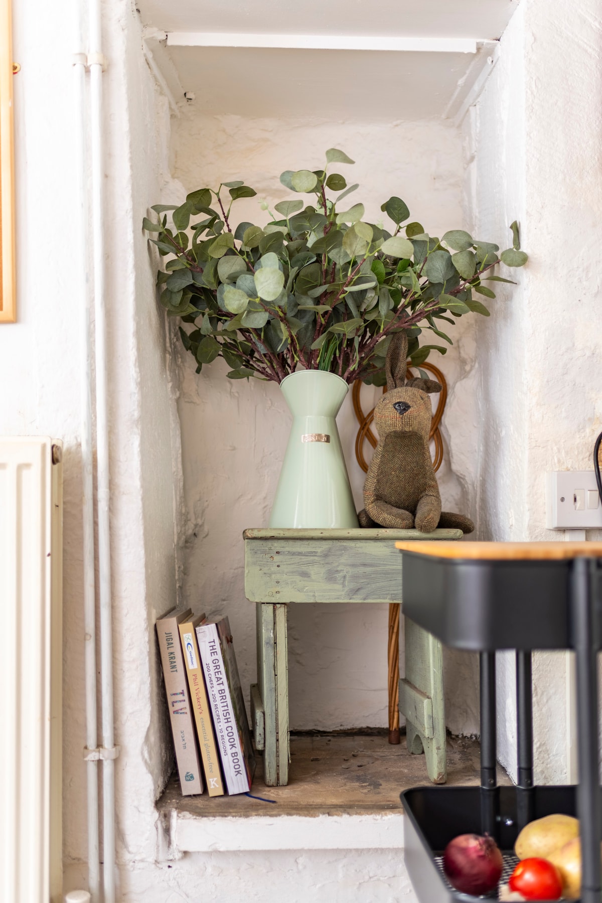 A small, green table is positioned against a white wall, adorned with a vase containing fresh eucalyptus. A plush rabbit toy sits nearby, adding a playful touch. Beneath the table, a few books are neatly stacked, enhancing the cozy ambiance of the space.