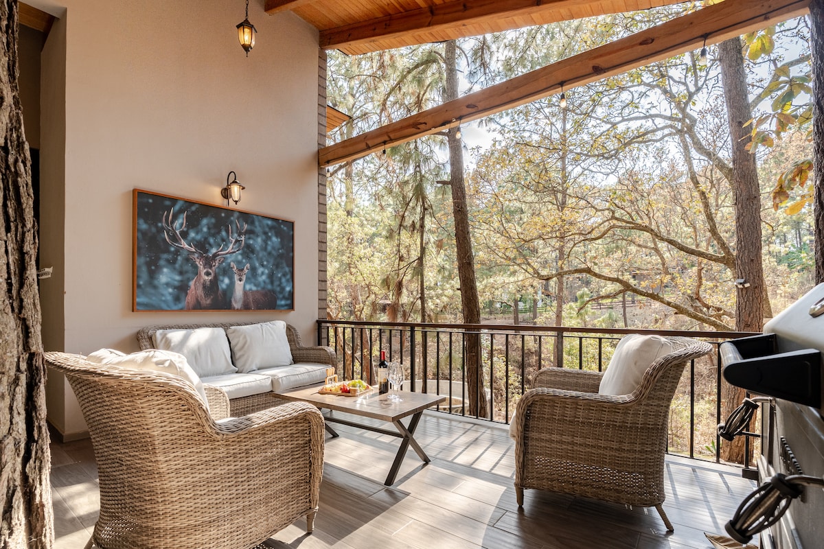 A covered outdoor seating area features woven chairs and a matching coffee table. A large artwork depicting deer adorns the wall. Trees with autumn foliage provide a natural backdrop, enhancing the peaceful ambiance of the space.