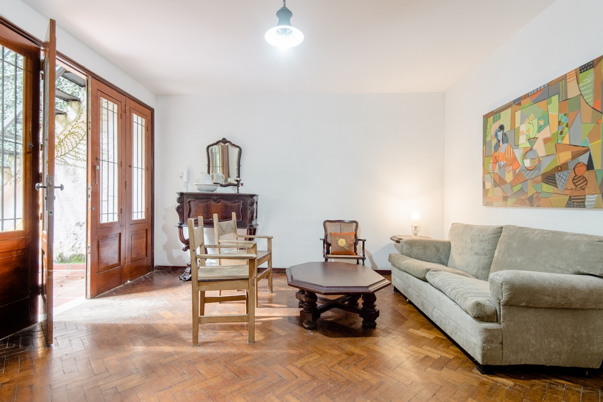 A spacious living area features large wooden doors that open to a garden view. Comfortable seating includes a light green sofa and four chairs surrounding a wooden coffee table. An art piece on the wall adds a touch of color, complementing the warm wood flooring.