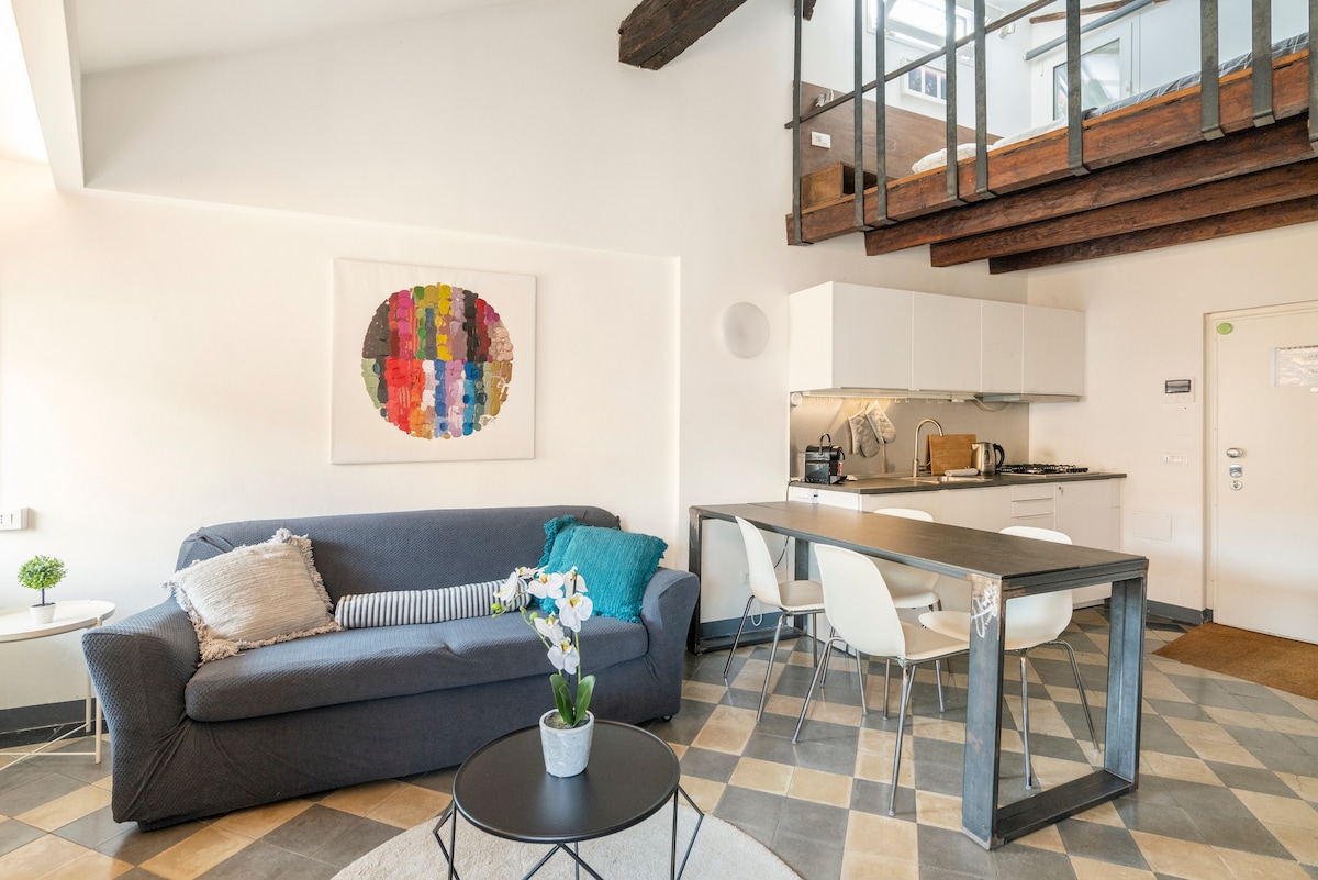 A spacious living area is highlighted by a comfortable sectional sofa and a round coffee table. A modern kitchen is visible with sleek cabinetry and essential appliances. The art piece on the wall adds a touch of color, and the tiled flooring enhances the room's ambiance.