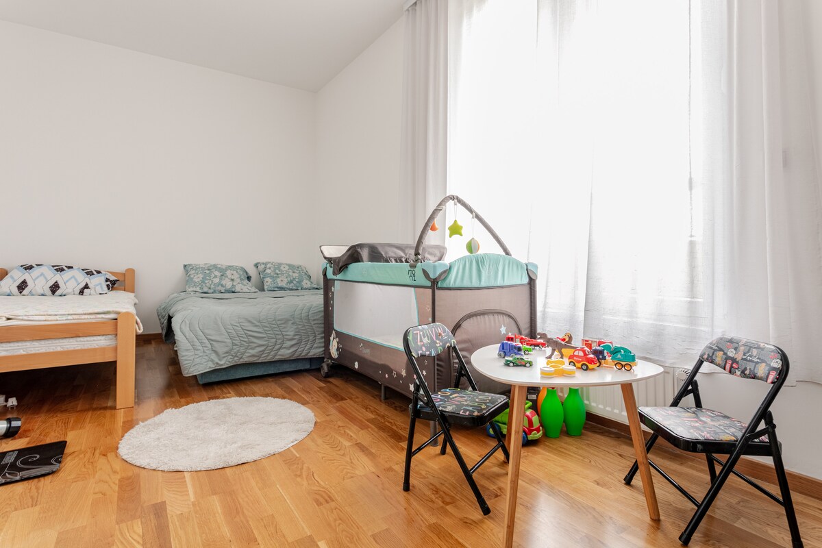 A spacious bedroom features a toddler bed alongside a single bed with light teal bedding. A portable crib is arranged near a window, with colorful toys placed on a small round table. Sunlight filters through sheer curtains, illuminating the warm wooden floor.