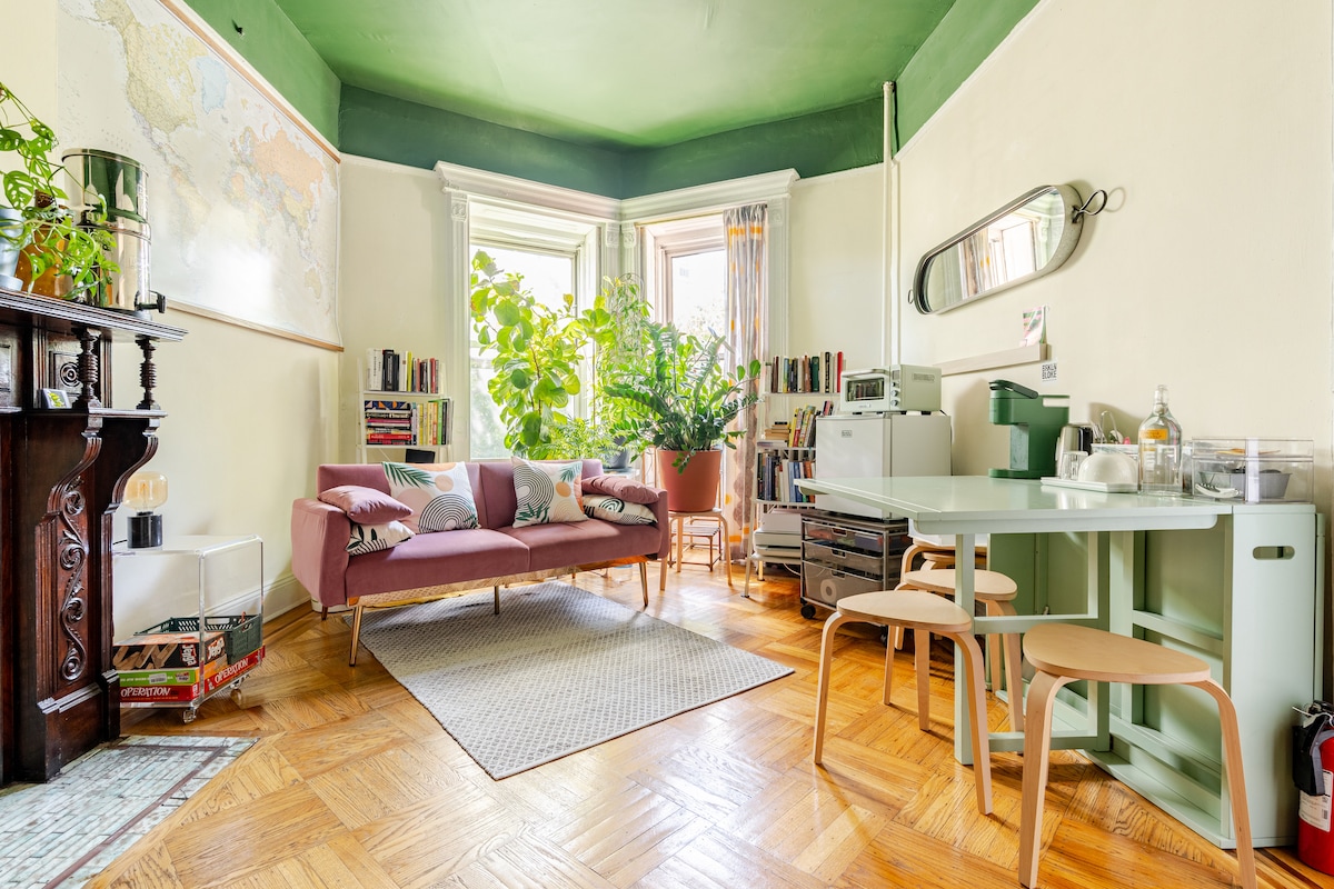 A shared living space features a pink sofa adorned with multiple cushions, surrounded by lush greenery. A dining table with light wooden chairs sits nearby. Natural light floods the room through large windows, highlighting the decorative plants and a world map on the wall.