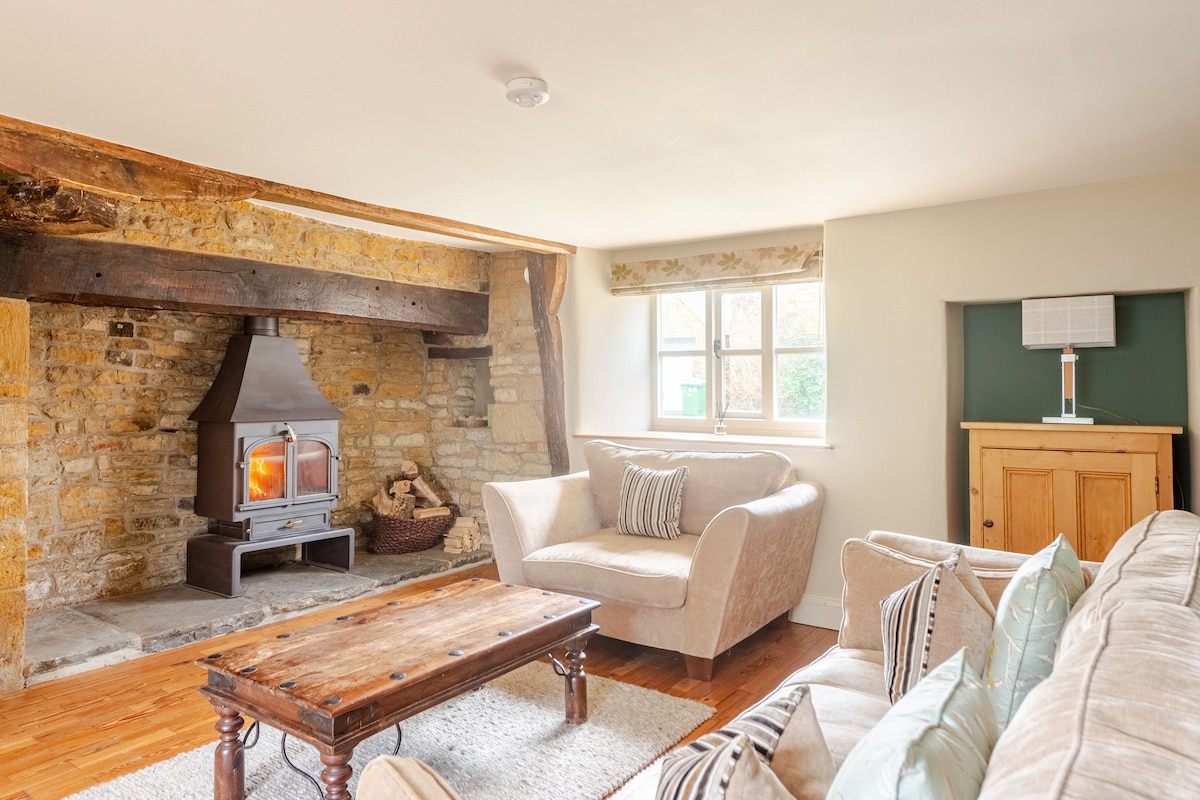 A cozy living area features a stone fireplace with a wood-burning stove, surrounded by exposed wooden beams. Two comfortable sofas face a wooden coffee table, while light enters through a window adorned with neutral-toned fabric. A wooden cabinet is positioned against the wall.