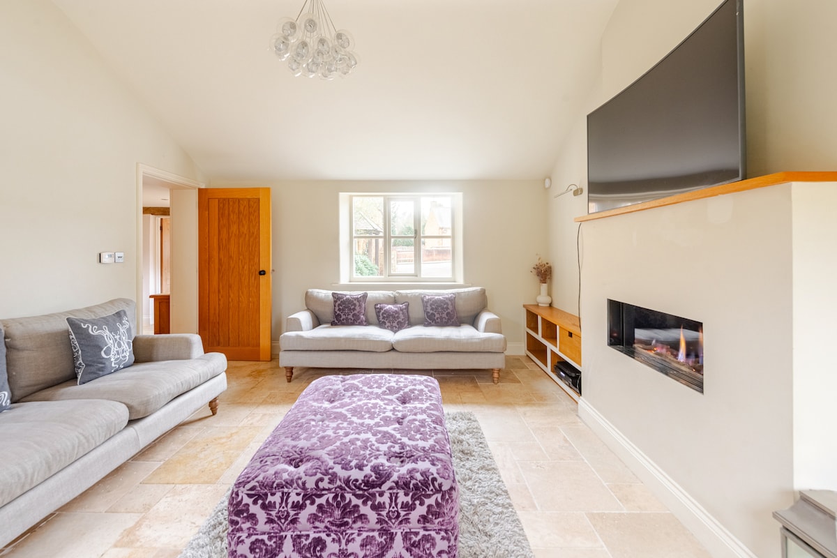 A spacious living area features a contemporary design with a large flat-screen television mounted above a modern fireplace. Comfortable seating is provided by a light-colored sofa and a plush, patterned ottoman, enhancing the cozy atmosphere of the room.