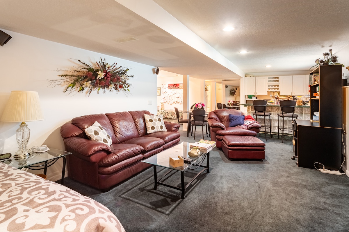 A spacious living area features comfortable leather seating arrangements, including a sectional sofa and armchairs. A glass coffee table occupies the center, while a kitchenette is visible in the background. Natural light illuminates the neutral-toned carpet, enhancing the welcoming ambiance.