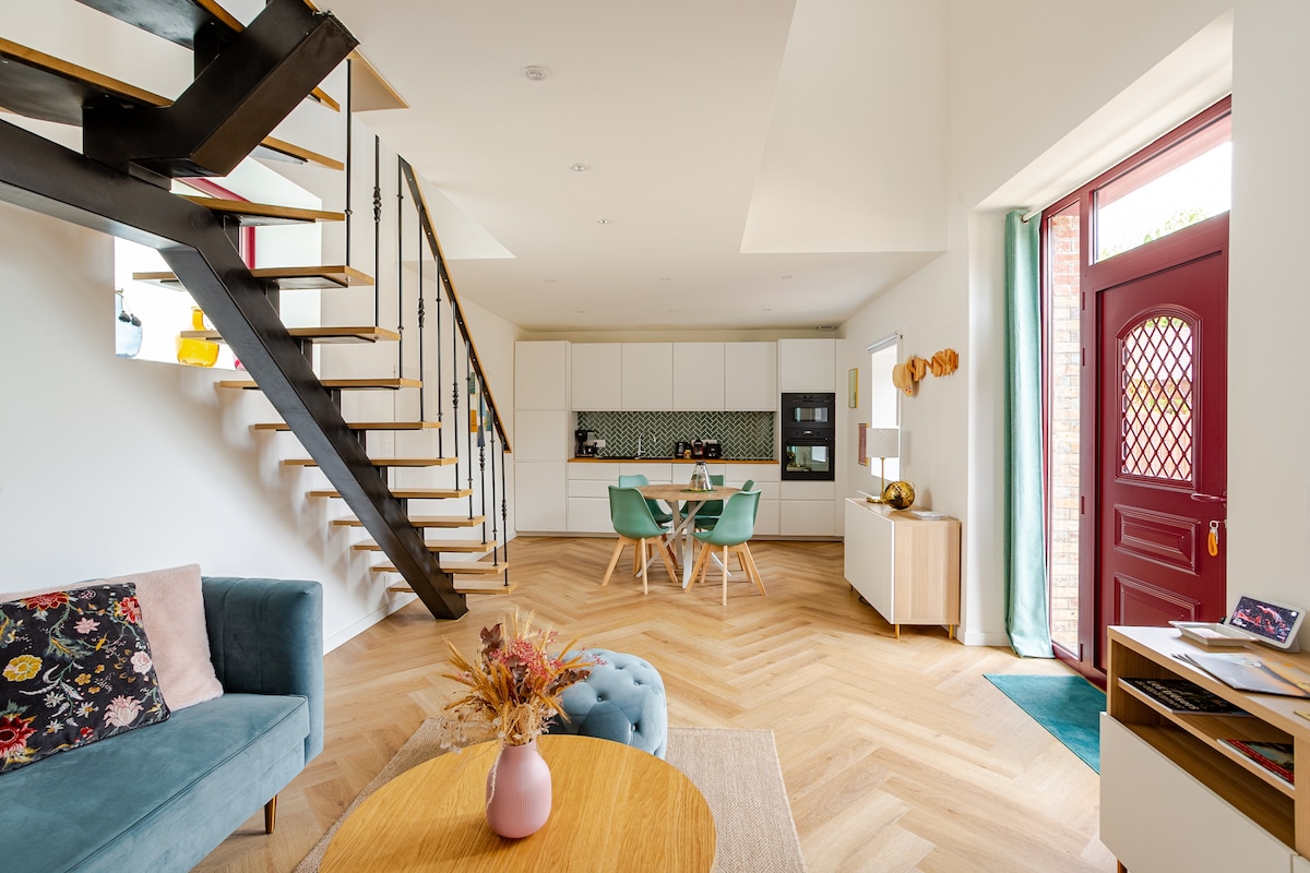 A spacious open-plan living area features a modern kitchen with white cabinetry and a patterned backsplash. A comfortable seating arrangement includes a blue sofa and a round coffee table. Natural light fills the space through large windows, while a distinctive staircase leads to the upper level.