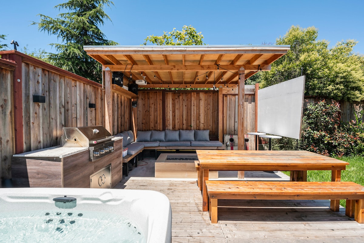 An outdoor entertainment area features a covered seating space with a sectional sofa and wooden tables. A barbecue grill is positioned nearby, alongside a projector screen. A hot tub is visible in the foreground, contributing to the inviting outdoor setting.