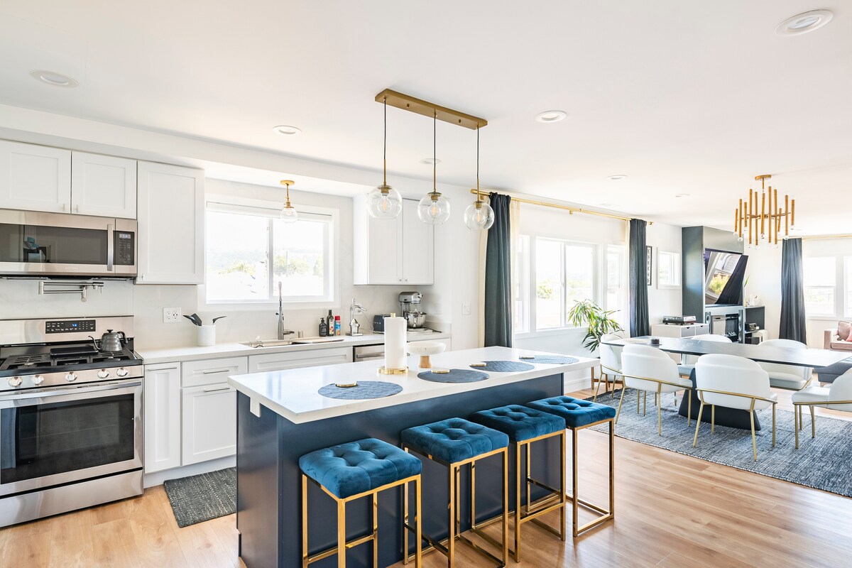 A modern kitchen features white cabinetry and stainless steel appliances, complemented by a large island with four blue upholstered stools. Natural light pours in through the window above the sink, illuminating the open layout that connects to a spacious living area.
