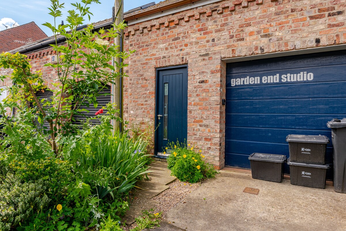 The entrance to the Studio is framed by lush greenery, featuring vibrant yellow flowers and dense foliage. A blue door with a clear nameplate leads into the property, set against a backdrop of textured brick walls. The adjacent area includes trash bins and a garage, emphasizing the residential setting.