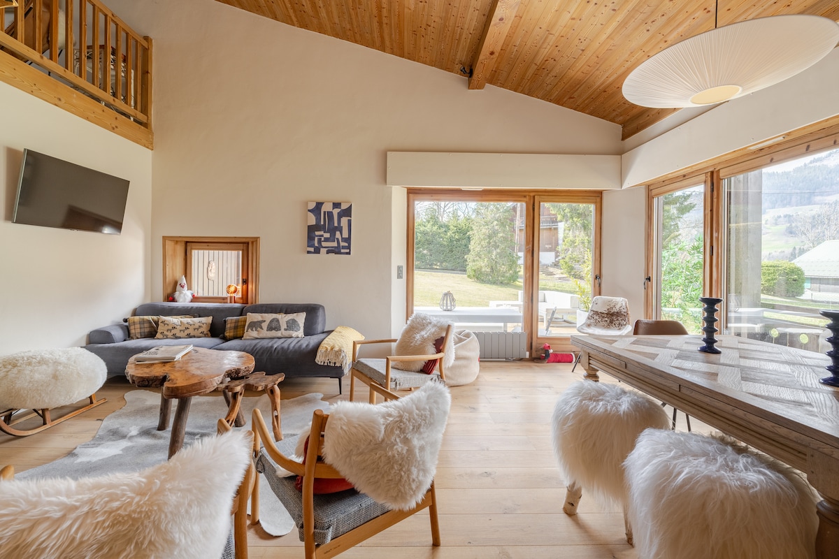 A cozy living space features a mixture of comfortable seating arrangements, including a couch and chairs with soft, textured throws. Large windows allow natural light to illuminate the room, showcasing views of the garden. A wooden table is positioned nearby, enhancing the warmth of the setting.