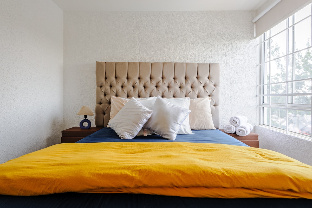 A stylish sleeping area features a tufted headboard with multiple pillows and a vibrant yellow blanket draped over the bed. A small lamp stands on a bedside table, while rolled towels are neatly arranged nearby. Natural light filters in through the window, enhancing the serene atmosphere.