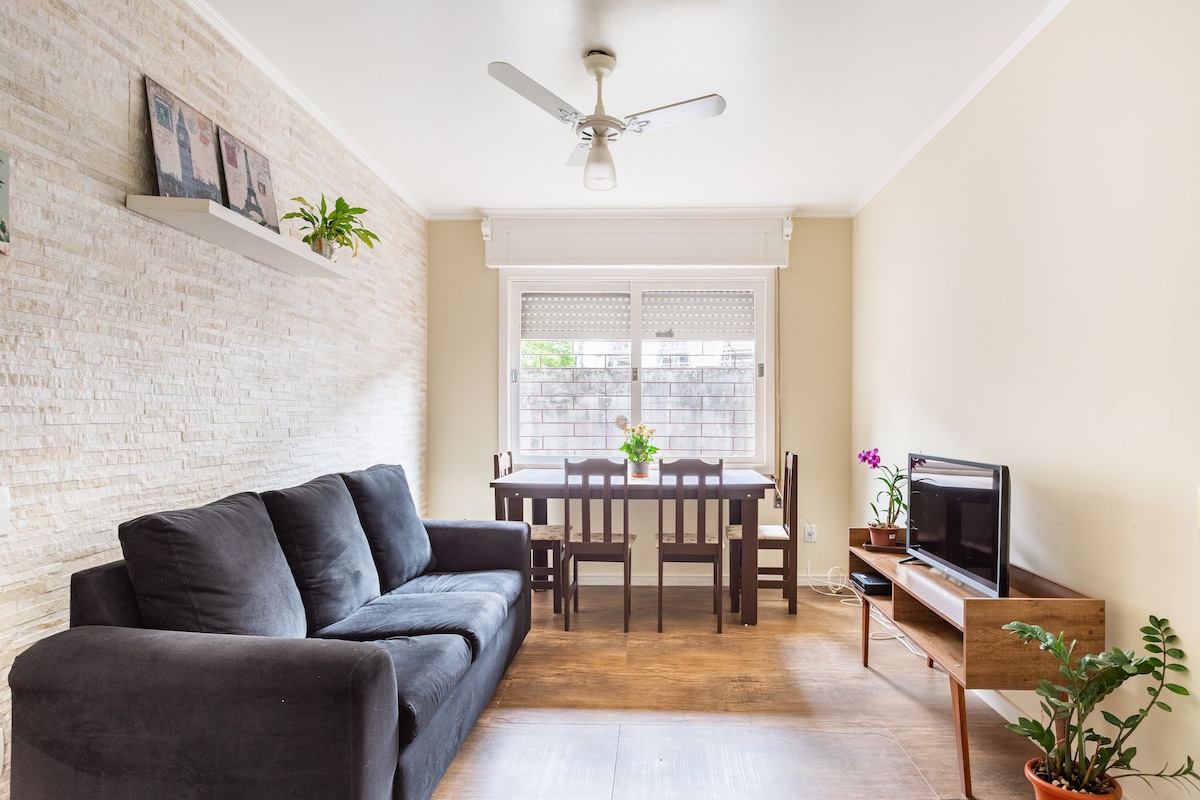 A cozy living area is visible, featuring a dark sofa and a wooden dining table surrounded by four chairs. Natural light enters through the window, illuminating a small indoor plant. Wall-mounted shelves display decorative items, while a television is positioned on a wooden stand.