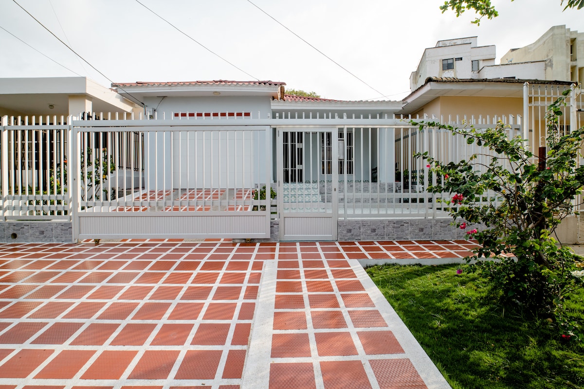 A secure and welcoming entrance is shown, featuring a white metal gate and a paved driveway. Green grass and flowering plants are visible alongside the path, leading to a modern exterior of the house, which has large windows and a covered porch area.