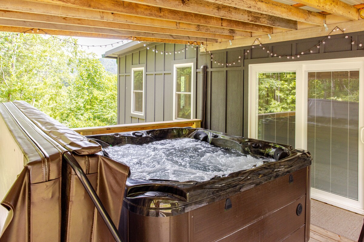 A hot tub sits on a covered deck, surrounded by wooden beams and outdoor string lights. Lush green trees are visible in the background through large sliding glass doors, allowing natural light to brighten the space.