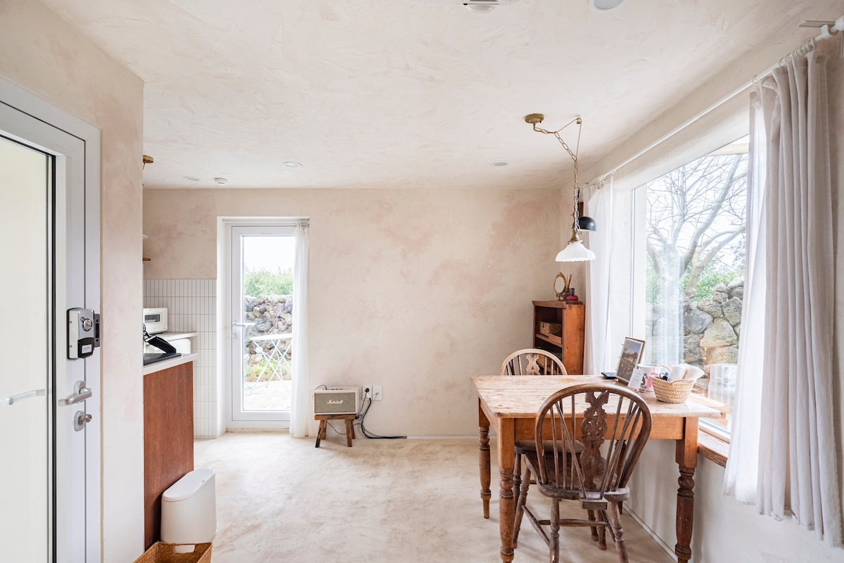 A cozy dining area features a wooden table surrounded by two chairs, illuminated by natural light streaming through two windows. A small kitchenette with a stove and sink is visible in the background, complemented by earthy, warm-toned walls.