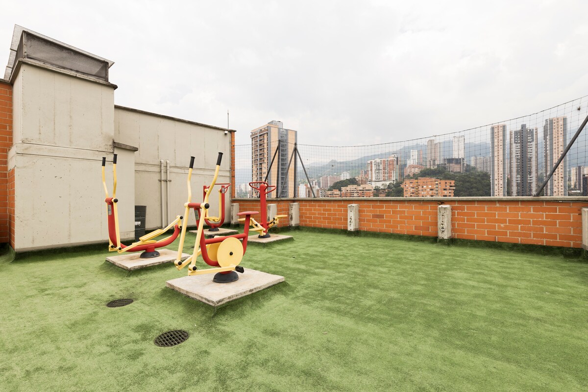 An outdoor exercise area is showcased, featuring several fitness machines positioned on a grassy surface. The backdrop reveals a panoramic view of the surrounding urban skyline, with a mix of tall buildings and distant mountains visible beyond.