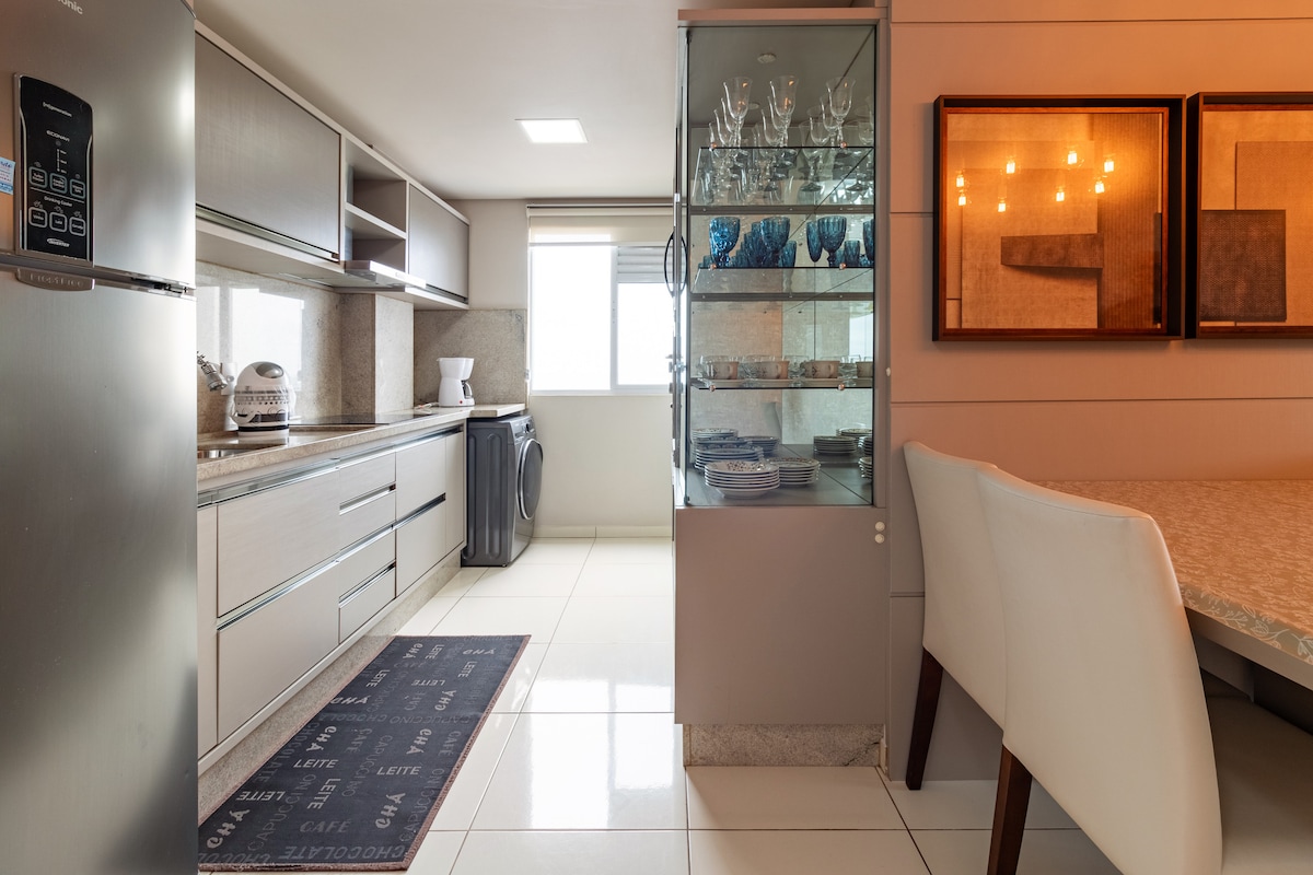 The kitchen area features modern cabinetry and countertops, with a stainless-steel refrigerator and a glass-fronted cabinet displaying glassware. A dining table with chairs is positioned near a window, allowing natural light to illuminate the space, complemented by a neutral color scheme.