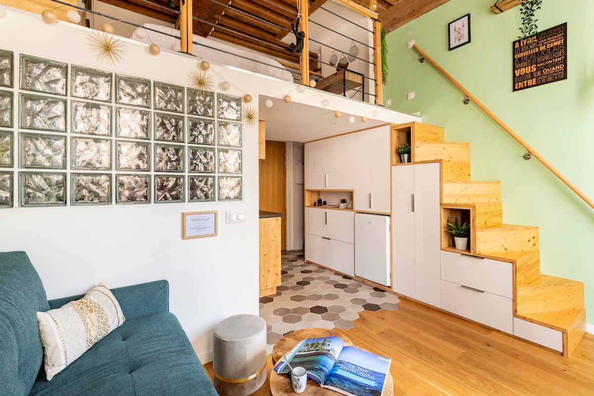 A modern living area features a comfortable green couch and a decorative wooden coffee table, with an open kitchen visible in the background. Wooden stairs lead to the mezzanine bedroom above. The wall displays a unique glass block design, enhancing the light-filled space.