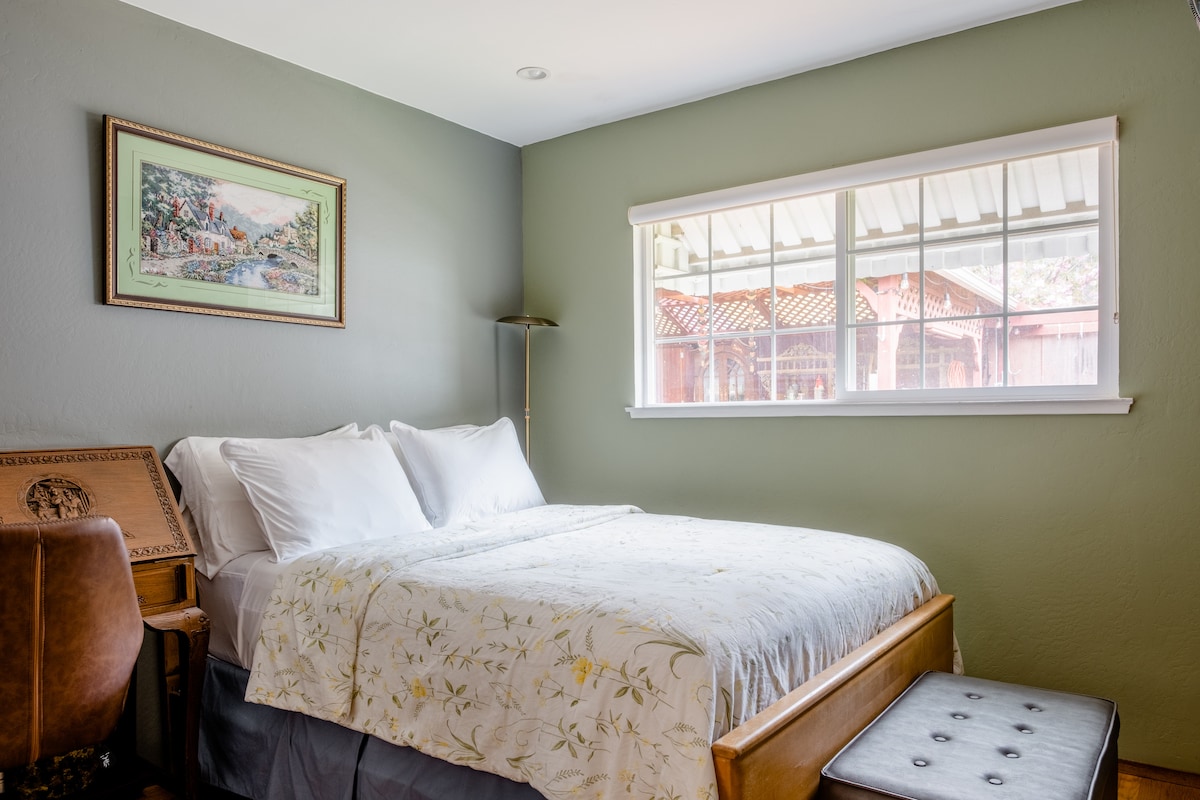 A well-lit room features muted green walls with a spacious bed dressed in white beddings and a floral-patterned comforter. Natural light flows through a wide window adorned with sheer curtains, creating a calm atmosphere. A Chinese writing desk is placed nearby.