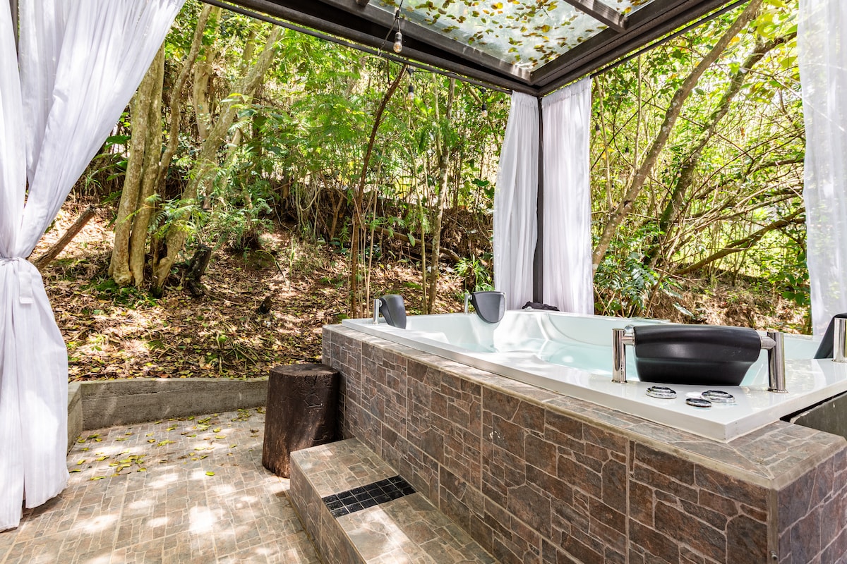 An outdoor hot tub is situated under a canopy with white curtains that billow gently. Surrounding greenery creates a serene backdrop, and the textured stone surface of the tub adds a natural element to the environment. Soft sunlight filters through the leaves above.