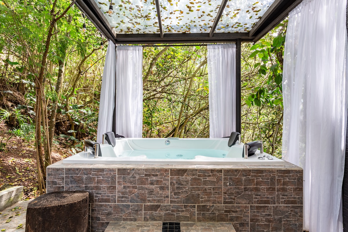 A relaxing hot tub is positioned beneath a glass canopy, surrounded by lush foliage. Sheer white curtains frame the space, creating an intimate atmosphere, while natural light filters through the canopy. The stone structure complements the serene woodland setting.