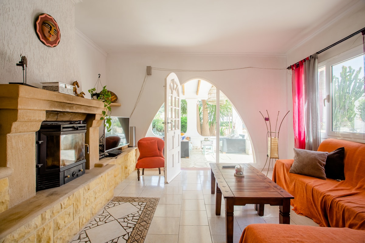 A spacious living room features an elegantly designed stone fireplace and a wooden coffee table. Two comfortable orange sofas invite relaxation, while large windows allow ample natural light. The room opens to an inviting outdoor area with patio space visible beyond the arched doorway.