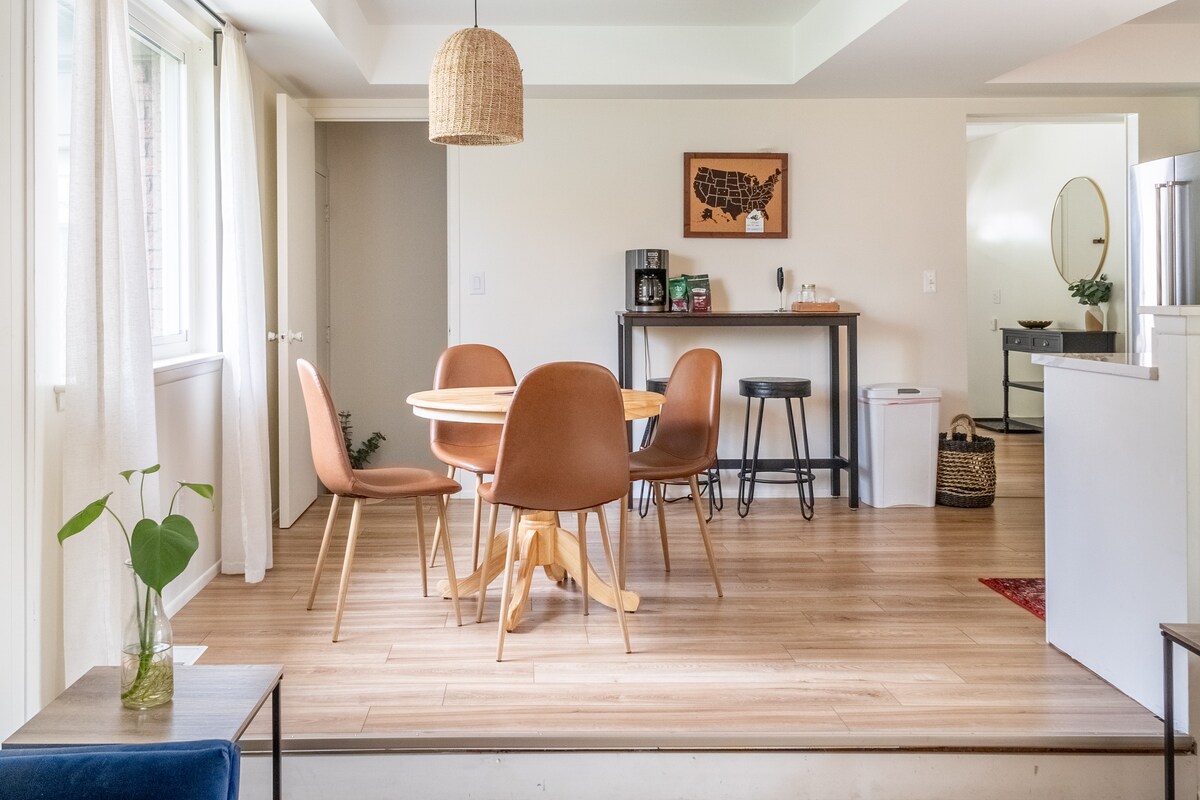 A bright, open dining area features a round wooden table surrounded by four modern chairs. Light pours in through large windows, illuminating the light-colored walls and wooden flooring. A small plant sits on a side table, adding a touch of greenery to the space.