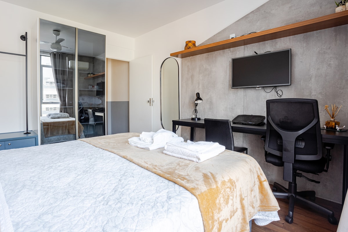 A bright bedroom features a spacious bed with a neutral-colored blanket. A desk and ergonomic chair are positioned next to a mounted television. Wardrobe doors and a mirror are visible, along with a ceiling fan above. Natural light filters through the window draped with light curtains.
