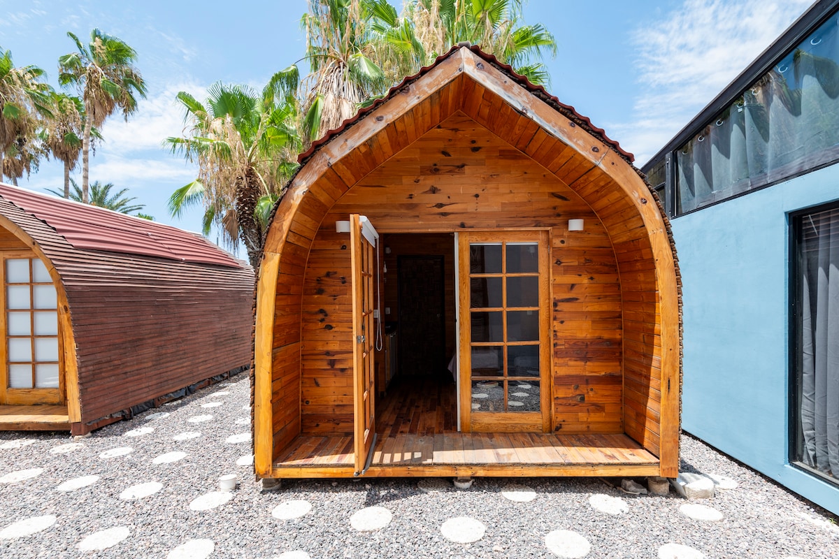 A rustic wooden cabin features a distinctive arched roof and double doors, inviting natural light into the interior. The surrounding area showcases textured gravel and lush palm trees, offering a serene outdoor environment for relaxation.