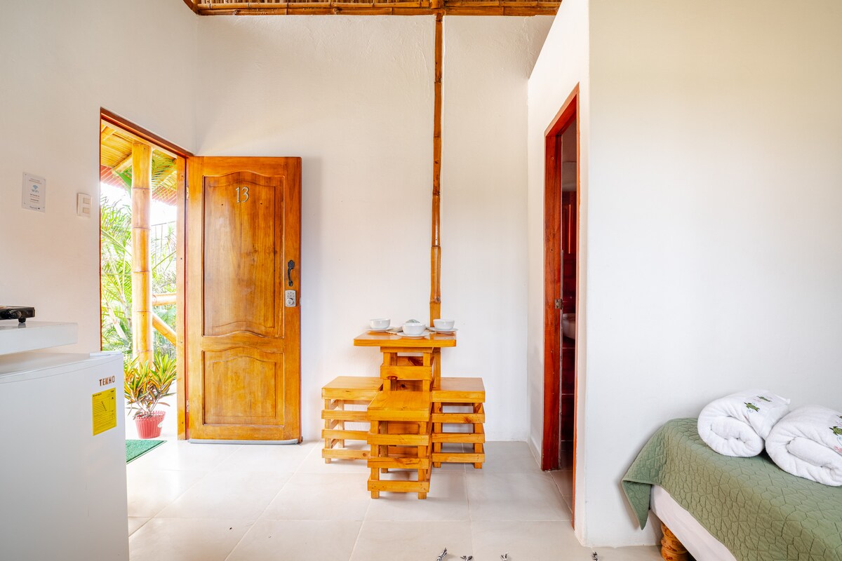 A welcoming entryway features a wooden door and a simple dining area with stacked wooden seating. Natural light illuminates the space, highlighting the bright walls and the greenery visible through the open door.