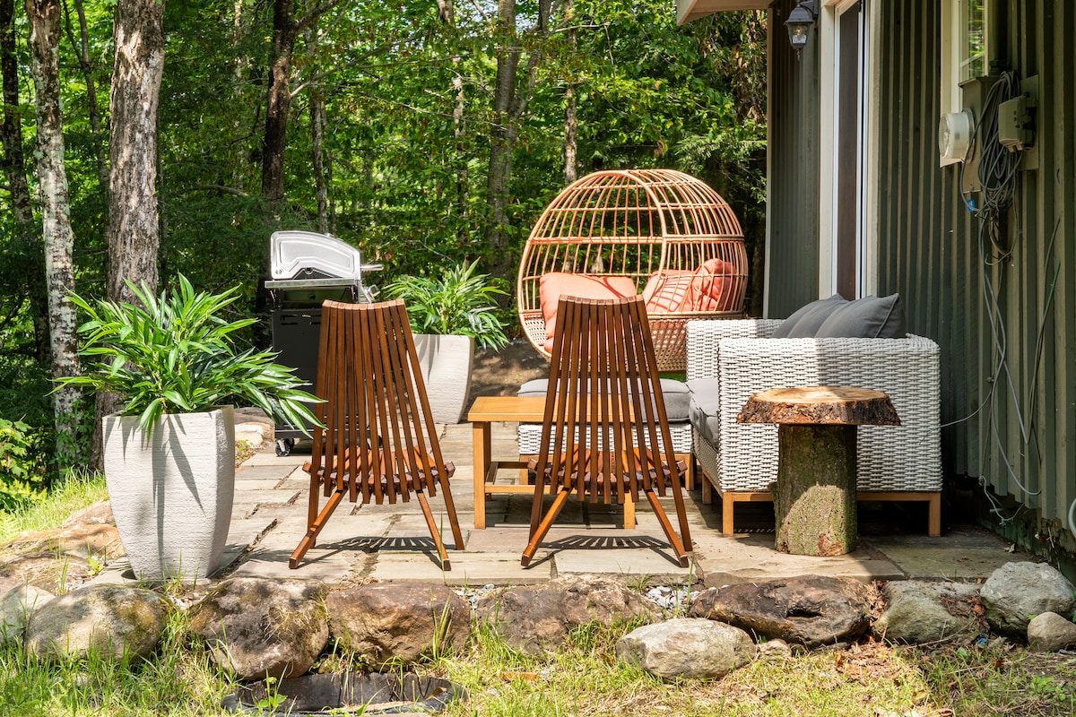 An outdoor patio area features two wooden lounge chairs facing a central table, surrounded by greenery. A cozy seating arrangement includes a modern sectional sofa and a distinctive round lounge chair. A barbecue grill is positioned nearby, with stone paving underfoot.