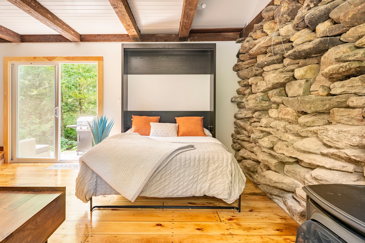 A spacious sleeping area is featured, highlighted by a Queen Murphy bed positioned against a natural stone accent wall. Large glass doors lead to the outside, allowing natural light to fill the room. Wooden flooring and simple decor contribute to a modern and serene ambience.