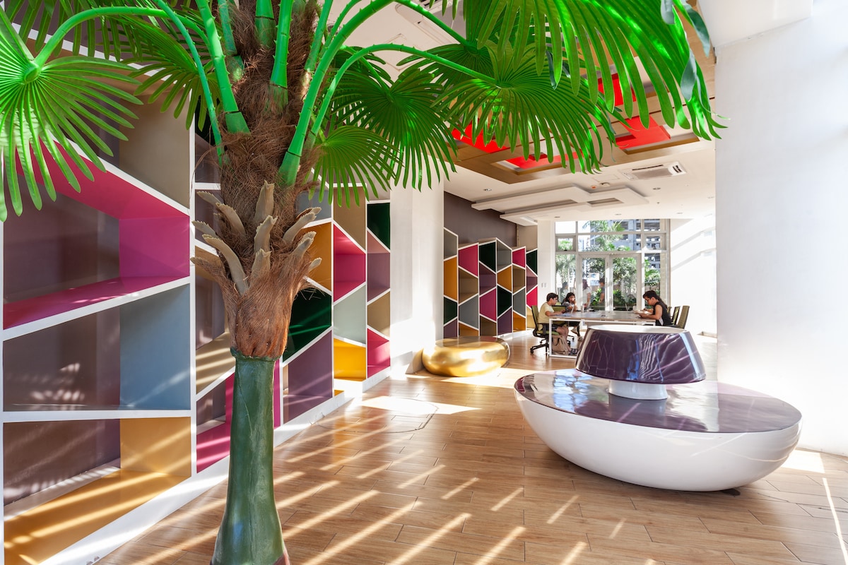 A vibrant common area showcases a large green artificial palm tree and colorful geometric shelving units. Natural light floods the space through expansive windows, illuminating a stylish white seating arrangement. Guests can be seen engaging in conversation, creating a lively atmosphere.