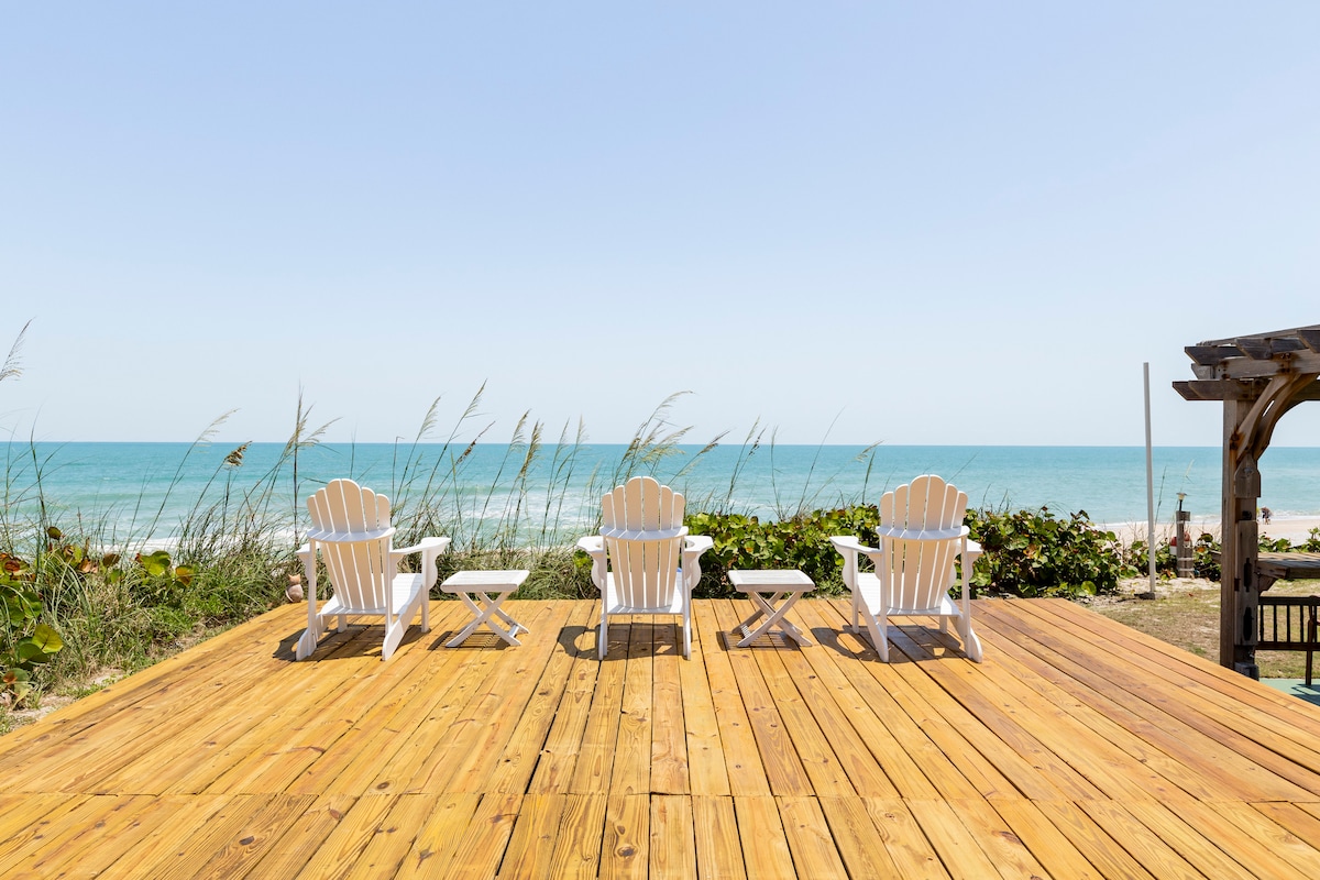 A spacious outdoor deck features four white Adirondack chairs facing the ocean. Light brown wooden planks create a warm surface, while gentle sea grass sways nearby. The calm blue water and clear sky create a serene backdrop, offering an inviting space to relax.