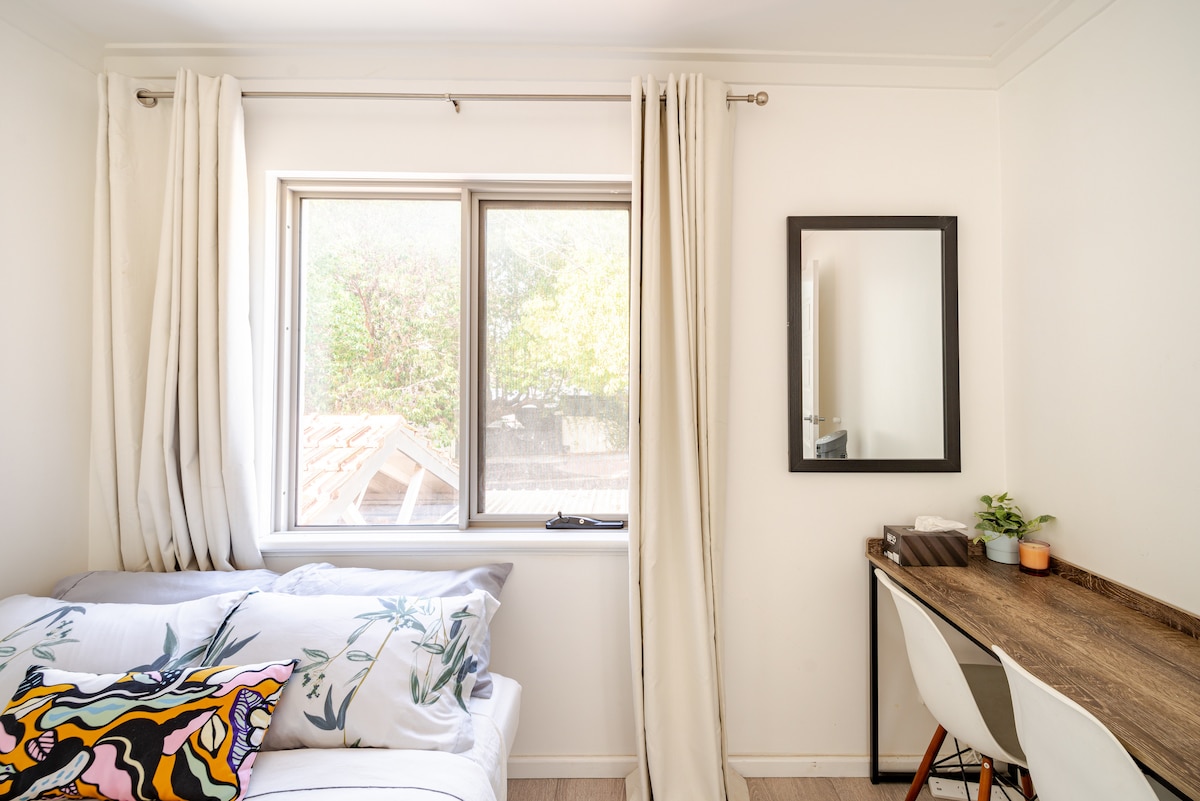 A bright room features a spacious bed adorned with floral-patterned pillows. Natural light enters through a large window, illuminating a simple wooden desk placed against the wall. A small mirror is mounted nearby, and a decorative plant adds a touch of greenery.
