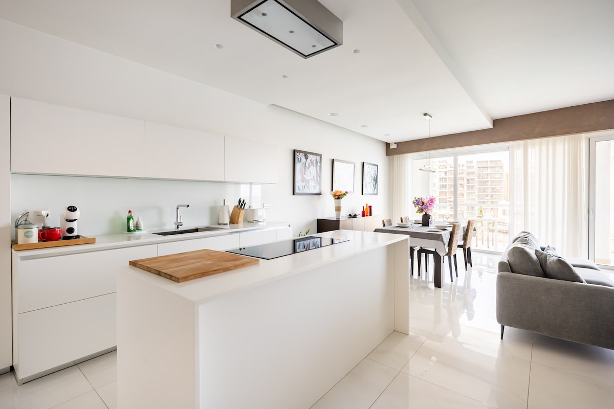 An open-plan kitchen and dining area is illuminated by natural light from floor-to-ceiling windows. The modern kitchen is equipped with sleek white cabinetry, an island with a built-in cooktop, and wooden cutting boards. A dining table with chairs is positioned nearby, complemented by a comfortable gray sofa.